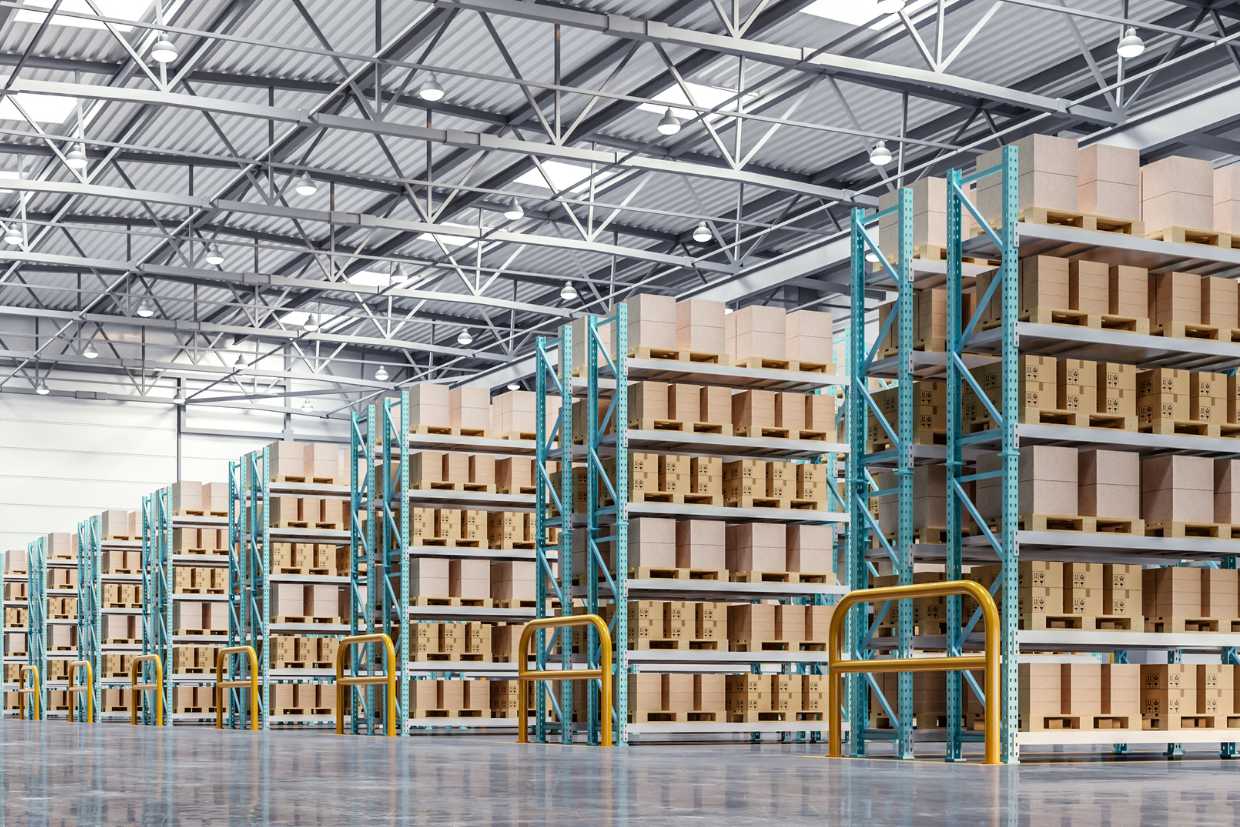 A spacious warehouse interior with multiple rows of tall shelving units filled with neatly stacked cardboard boxes. The warehouse has high ceilings with visible metal beams and bright overhead lighting.