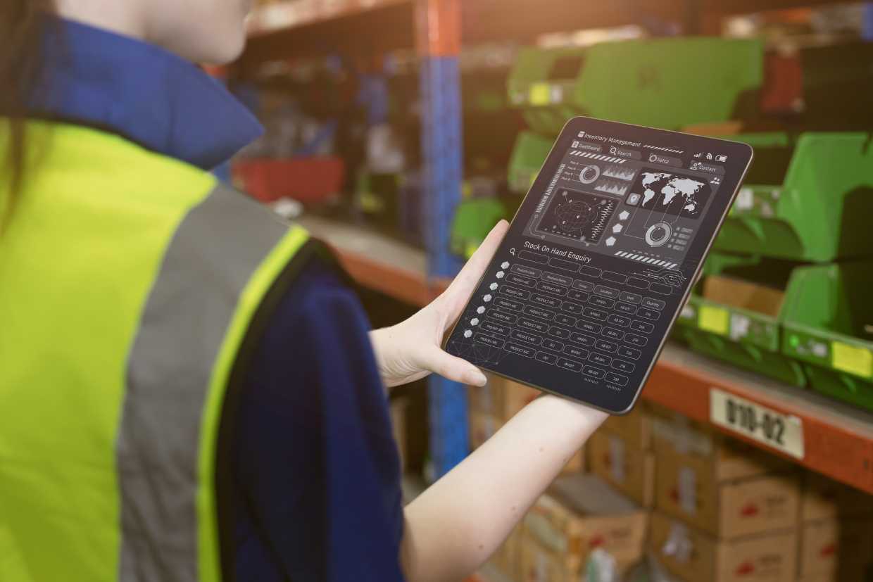 A person wearing a high-visibility vest holds a tablet displaying an inventory management interface in a warehouse setting. Shelves with various items are visible in the background, indicating an organized storage area.