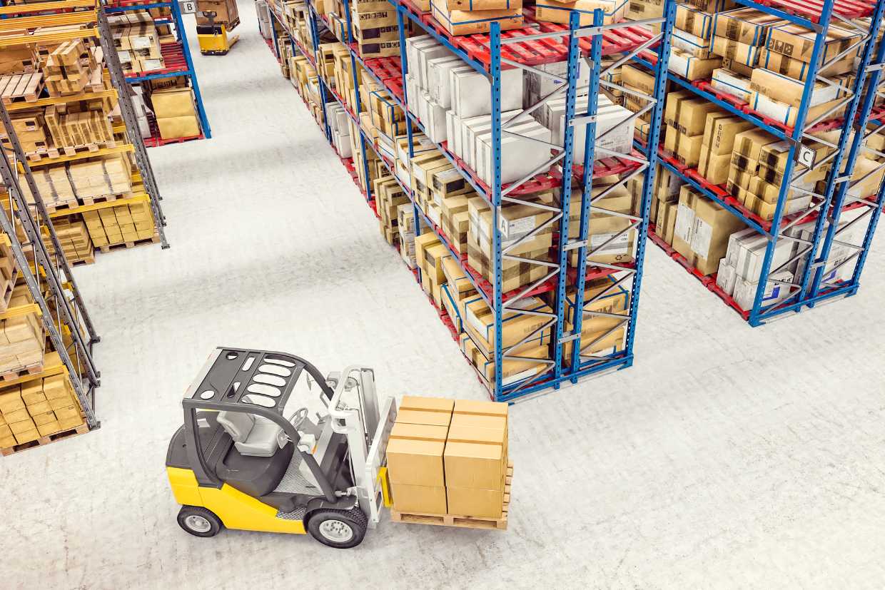 A forklift transporting a pallet with stacked boxes in a warehouse. The warehouse has multiple rows of tall shelving units filled with various items, demonstrating an organized and efficient storage system.