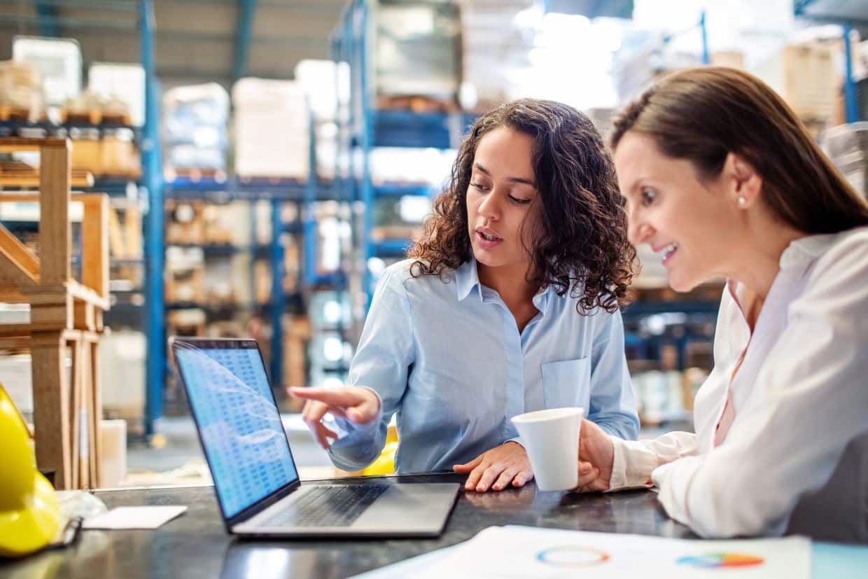 Two women in a warehouse discussing data on a laptop screen. One is pointing at the screen while explaining something, and the other is smiling while holding a coffee cup. Shelving with stocked inventory is visible in the background, indicating a warehouse management setting.