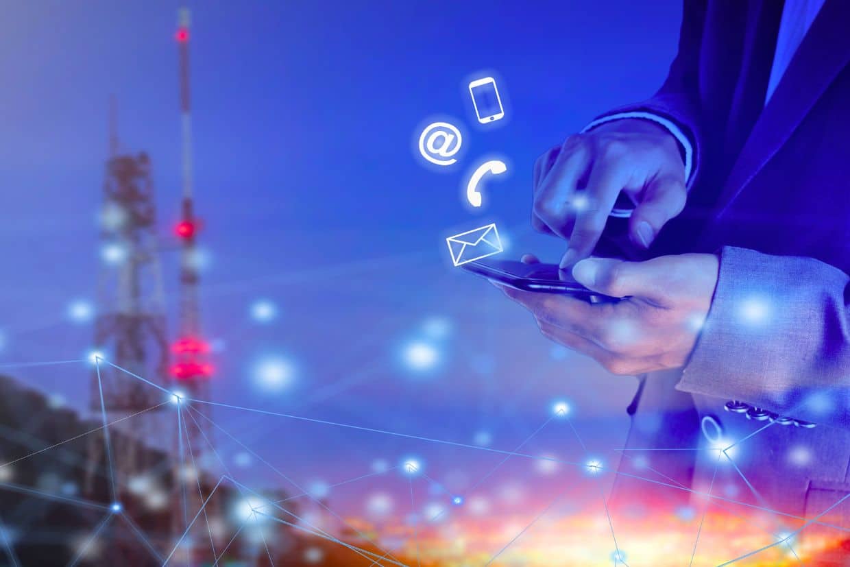 A close-up of a hand holding a mobile phone, with glowing icons representing email, phone calls, and location hovering above the device. A radio tower with red lights and a blurred cityscape at night are visible in the background, suggesting connectivity and communication networks.