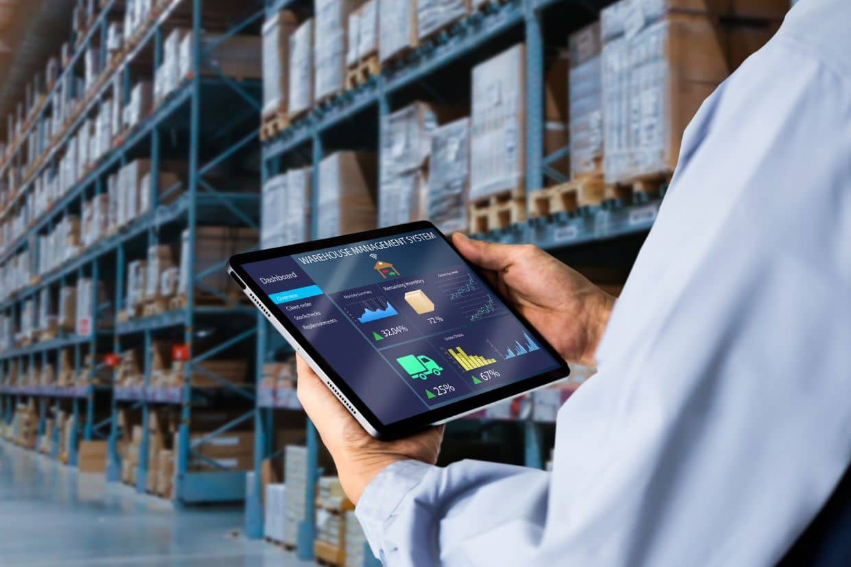 A warehouse worker uses a tablet displaying a warehouse management system dashboard.