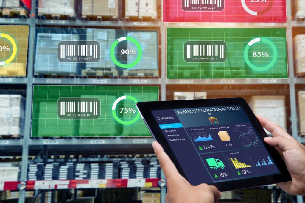 A person holding a tablet displaying a warehouse management system (WMS) dashboard with various metrics and graphs, including inventory levels, order status, and shipping details. In the background, shelves stocked with boxes are visible, each labeled with barcodes and percentage indicators showing inventory status.